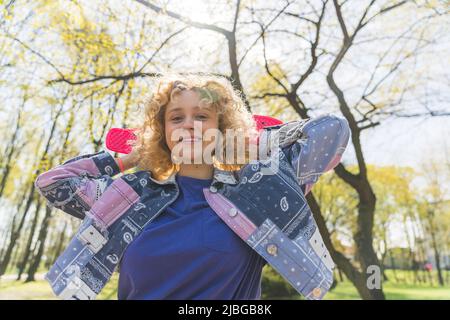 Horizontal outdoor shot of a pretty young joyful girl with blonde hair standing in the sun looking at a camera holding a pink small skateboard behind her head. High quality photo Stock Photo