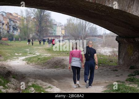 Couple Walking on the Lawn along the Banks of a River and a Bridge and Houses in the Background. Stock Photo