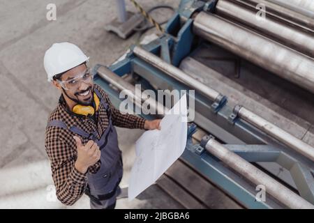 High angle view of heavy industry worker with safety headphones and hard hat in industrial factory holding blueprints Stock Photo