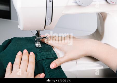 Woman tailor works on sewing machine. Hands hold fabric. World tailors day. Close-up of sewing process. Home clothing repair. Stock Photo