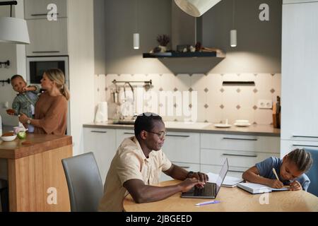 Lifestyle scene of multiethnic family with two kids and black father using laptop in foreground, copy space Stock Photo