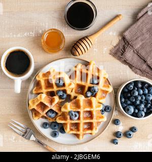 Sweet belgian waffles with blueberries, honey and cup of black coffee on a wooden table, top view. Square crop Stock Photo