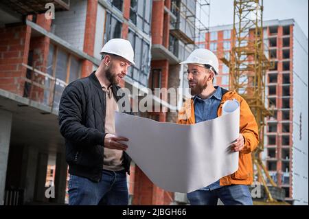 Bearded man in safety helmet holding architectural plan and talking with construction worker outdoors. Male homeowner discussion building plan with specialist at construction site. Stock Photo