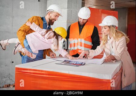 Woman standing next to builder and pointing at building plan while man holding daughter. Family with child discussing architectural plan of new apartment with construction worker. Stock Photo