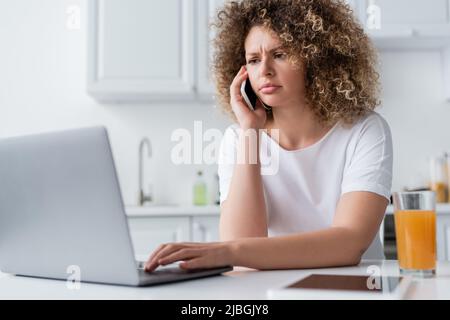 serious and frowning woman using laptop and talking on cellphone in kitchen Stock Photo