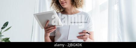 partial view of curly woman holding digital tablet and notepad, banner Stock Photo