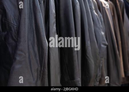 Dark leather jackets hang side by side in an open air market and are lit by the sun. Focus on the left jacket. Background image Stock Photo