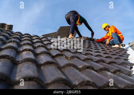 2 roofers working on the working at height to install the Concrete Roof Tiles on the new roof of new modern building construction. Stock Photo