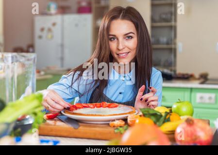 Pretty young woman smiling at camera while decorating a cake with sliced strawberry sitting in big kitchen. Stock Photo