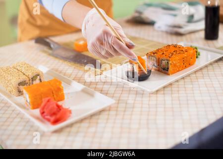 Close-up image of female hand glove holding a sushi roll in chopsticks dipping it in soy sauce in kitchen. Stock Photo