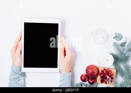 Tablet pc template with blank screen in female hands next to gift box, decorations and Christmas tree branches on white table, top view. Stock Photo