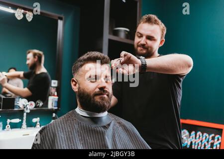 Professional barber doing haircut to bearded male customer in barbershop Stock Photo