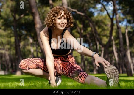 Cute caucasian woman wearing black sports bra standing on city park, outdoors stretching leg muscles, doing side lunges in skandasana pose. Healthy li Stock Photo