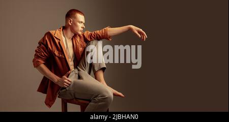 Portrait of brutal young red-haired man, sitting on chair, posing isolated over brown background. Flyer Stock Photo