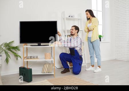Young friendly service man installs and connects new TV in female client's house. Stock Photo