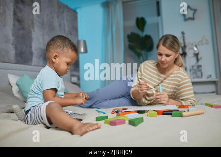 Portrait of cute black toddler playing with building blocks on bed, Caucasian mother in background, copy space Stock Photo