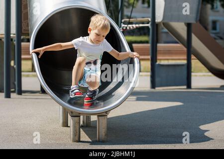 Boy child climbing sliding and playing on metal slide on outdoor playground in summer park. Activity and amusement center in kindergarten or school ya Stock Photo