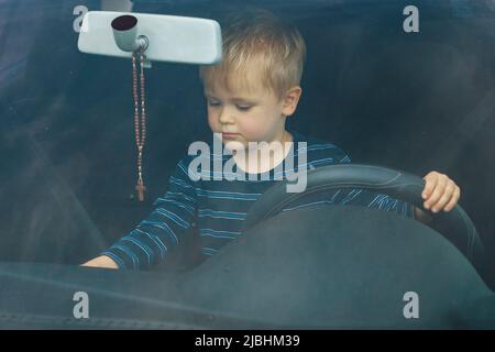 Cute little boy driving fathers car. Portrait of a child sitting in a car behind the wheel from the front. The boy plays and imagines he is driving, h Stock Photo