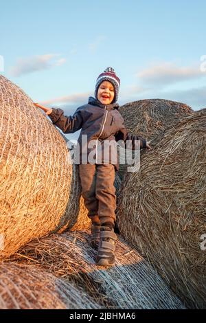 A smiling cute boy with a knitted hat stands on hay bales in the evening sunlight. Stock Photo
