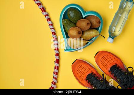 Orange sports sneakers, a gymnastic hoop wrapped in a measuring tape with a meter, a plastic bottle of water and a heart-shaped bowl filled with fruit Stock Photo