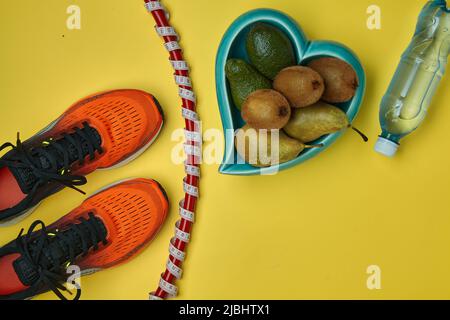 Orange sports sneakers, a gymnastic hoop wrapped in a measuring tape with a meter, a plastic bottle of water and a heart-shaped bowl filled with fruit Stock Photo