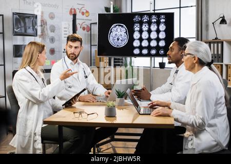 International medical team having meeting in conference room at modern hospital. Doctors discussing and interacting each other in laboratory with display showing brain MRI image on background. Stock Photo