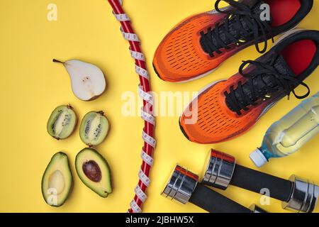 Orange sports sneakers, a gymnastic ring wrapped in a measuring tape with a meter, dumbbells, a bottle of drinking water and halves of cut fruit on a Stock Photo