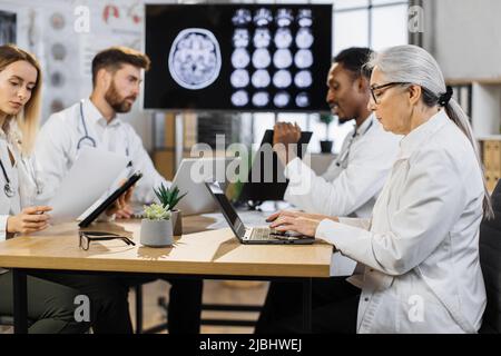 Portrait of senior female doctor sitting at desk with laptop. Group of multiethnic physicians taking online notes on different gadgets to patient history near. Stock Photo