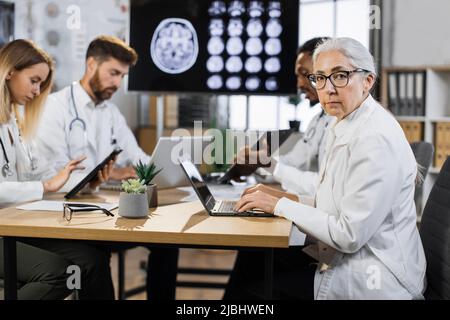 Portrait of senior female doctor sitting at desk with laptop and looking at camera. Group of multiethnic physicians taking online notes on different gadgets to patient history near. Stock Photo