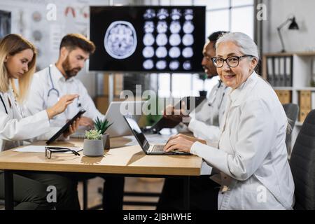 Portrait of senior female doctor sitting at desk with laptop and looking at camera with thumb up. Group of multiethnic physicians taking online notes on different gadgets to patient history near. Stock Photo