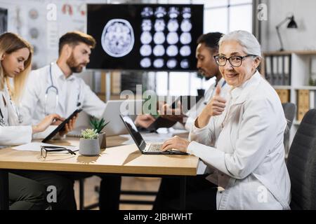 Portrait of senior female doctor sitting at desk with laptop and looking at camera. Group of multiethnic physicians taking online notes on different gadgets to patient history near. Stock Photo