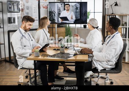 Team of multi ethnic medical researchers gathering at boardroom for having online briefing with male head doctor. Qualified therapists in lab coats using modern gadgets at hospital. Stock Photo