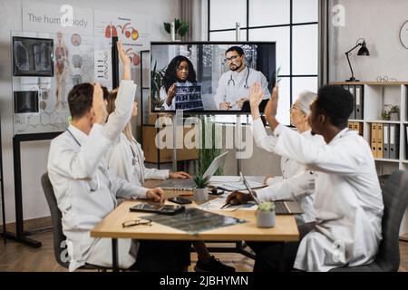 Team of multiethnic healthcare doctors sitting at table with arms raised during online video meeting. Medical workers in white lab coats voting for new successful tomographic project in modern clinic. Stock Photo