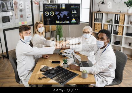 Top view of multiethnic hospital colleagues in face masks giving fist bumps for sharing positive mood. Doctors using modern digital devices at morning staff meeting during covid 19 pandemic. Stock Photo