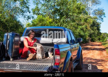 A woman sitting in the back of a pickup truck with her dogs and suitcase, all on a typical red sand road in Paraguay. Stock Photo