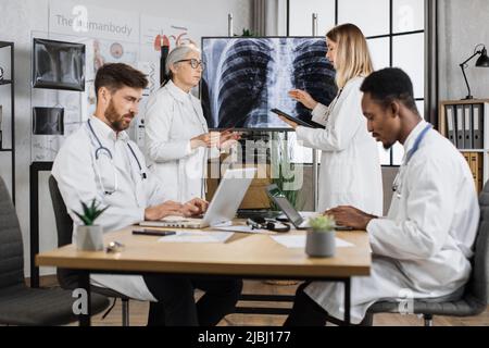 Group of two different aged female doctors talking about x ray results while multiethnic males working on laptops. Competent employees giving advices and sharing knowledge in modern clinic boardroom. Stock Photo