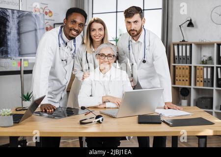 Happy researchers posing on camera with smile in front of laptop while working on new scientific project. Multiethnic lab staff making medical discoveries in modern pharmaceutical company. Stock Photo