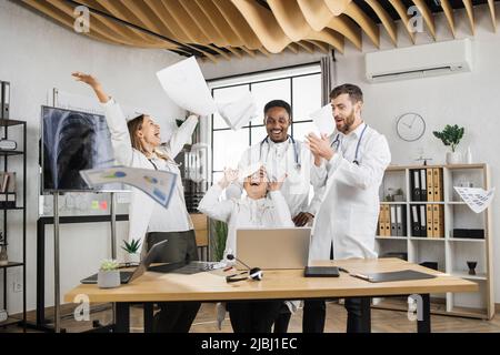 Excited multiracial scientists in white medical uniform laughing and throwing documents in air during laboratory meeting. Success teamwork and healthcare achievement concept. Stock Photo