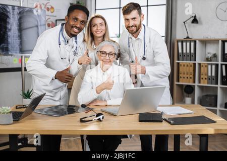 Happy researchers posing on camera with smile in front of laptop while working on new scientific project. Multiethnic lab staff making medical discoveries in modern pharmaceutical company withthumb up Stock Photo