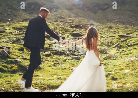 Bride in a dress with lace and groom holding hands and walking during their travel through the mountains. Nature landscape. Stock Photo