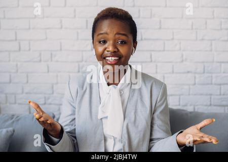 Smiling african woman having video call talking with co-workers or students, gesturing at web camera while sitting on the sofa. Online education Stock Photo