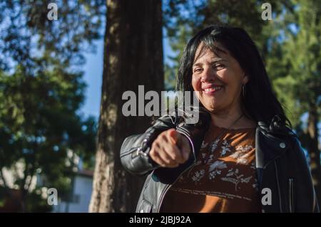 portrait latin caucasian adult woman, outdoors in countryside, standing smiling and pointing finger at camera, copy space to left. Stock Photo