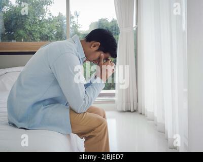 Thoughtful Asian man in denim shirt sitting on the bed with hands touching on head feeling lonely, looking down with sadness and thinking in the bedro Stock Photo