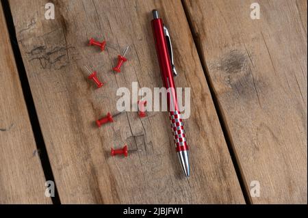 A red pen and a push pin are against the wooden background. A metal ballpoint pen and six push pins lie on top of old cracked boards. Selective focus. Stock Photo