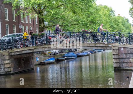 Amsterdam, Netherlands May 21, 2022. People bike on a bridge, over canal water, parked bicycles, moored boats. Traditional houses at Holland capital Stock Photo
