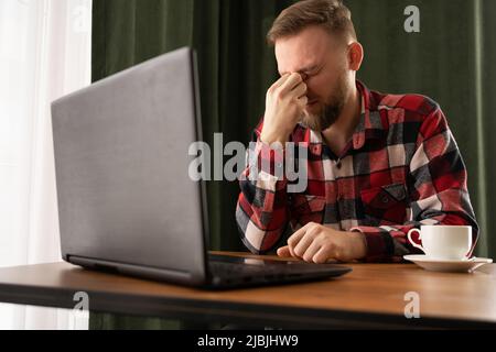Eye fatigue. Tired caucasian guy massaging the nasal bridge after working on a laptop computer while sitting at the workplace in the home office. Poor Stock Photo