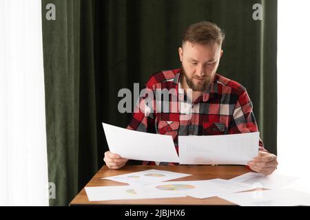 Young businessman working from home, holding a sketch in his hands, working with paper documents, remote analyst analyzes the work of employees Stock Photo