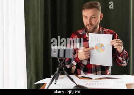 Young man making business presentation and demonstrating graphs, explaining charts to colleagues while video calling via smartphone Stock Photo