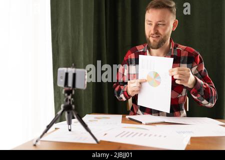 Caucasian young analyst manager making video call with colleagues presenting new project using smartphone and paper charts, video conferencing. people Stock Photo