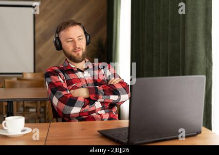 Pensive bearded man in headphones sitting on publicity area with laptop computer and listening audio book about modern technology, thoughtful man Stock Photo
